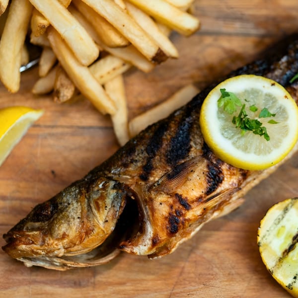 Grilled whole fish with lemon slices and parsley, served with golden french fries on a wooden board.