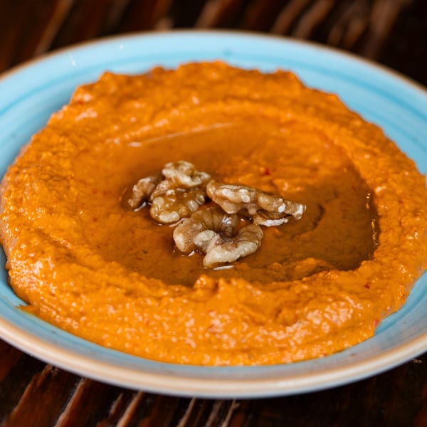 Bright orange muhammara dip, drizzled with olive oil and garnished with walnuts, in a blue bowl.