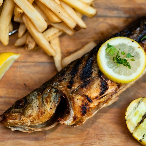Grilled whole fish with lemon slices and parsley, served with golden french fries on a wooden board.