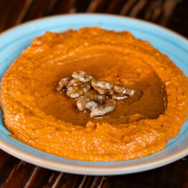 A vibrant orange Mediterranean dip, likely muhammara, garnished with walnuts and olive oil in a blue bowl.