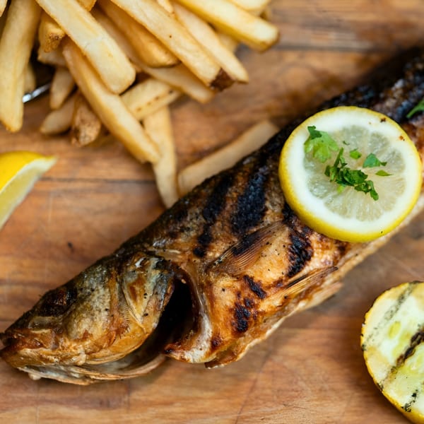 Grilled whole fish with lemon slices and parsley, served with golden french fries on a wooden board.