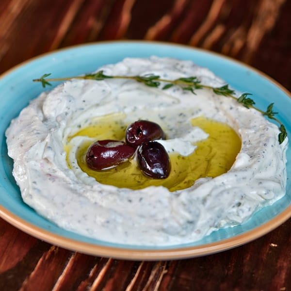 Creamy Mediterranean dip with black olives, olive oil, and a sprig of thyme in a blue bowl.