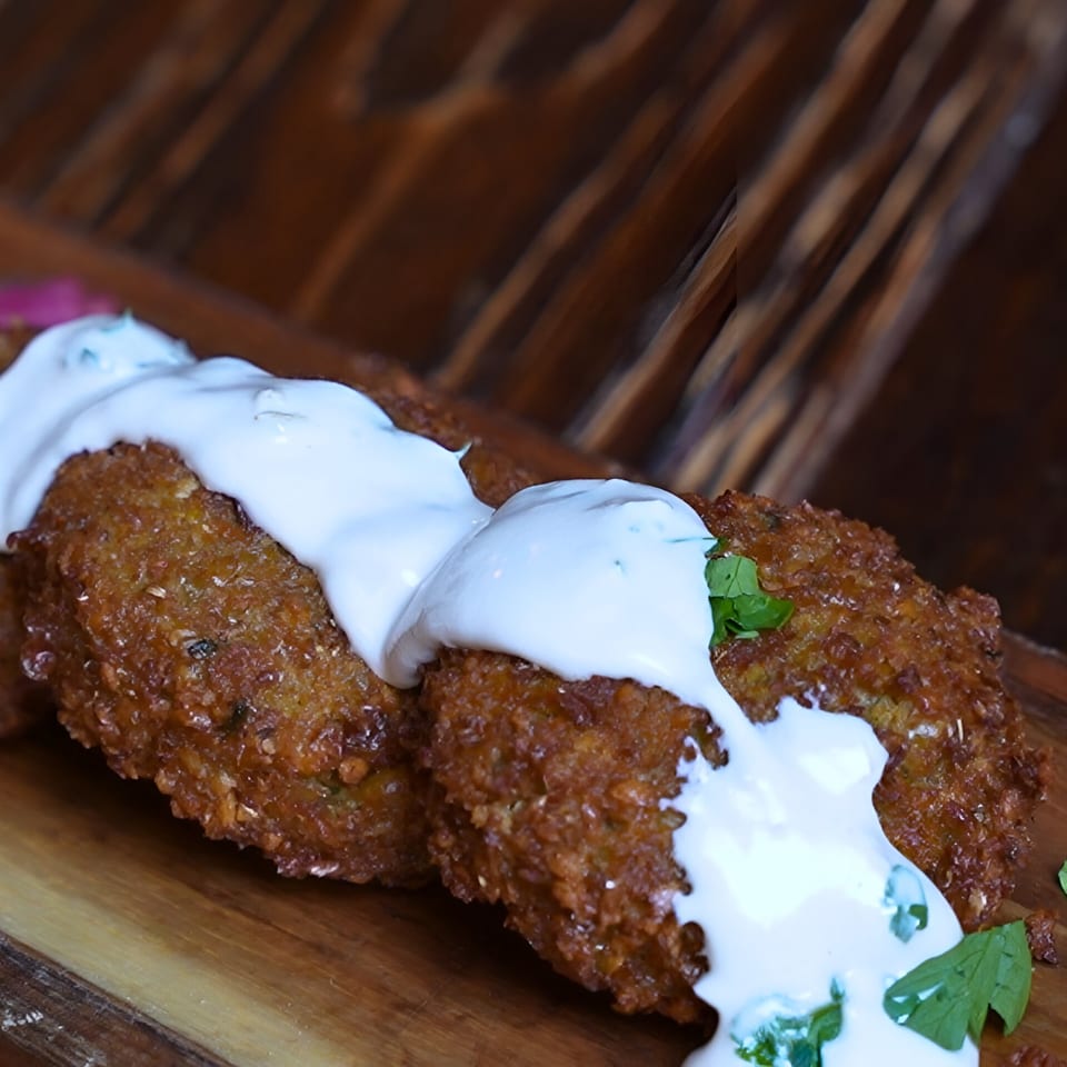 Crispy falafel patties drizzled with white sauce and garnished with fresh herbs on a wooden board.